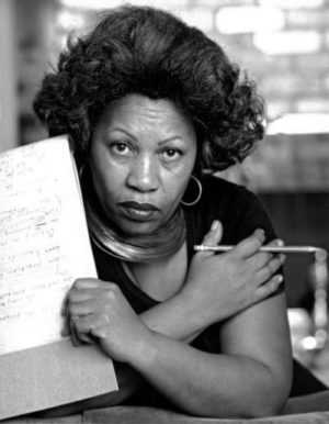 a black and white picture of Toni Morrison holding a piece of paper in one hand and a pencil in the other. her arms are crossed over her chest and she is staring directly at the camera.